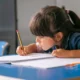 Menina concentrada escrevendo em um caderno com um lápis amarelo, rodeada de canetas coloridas em um ambiente escolar.