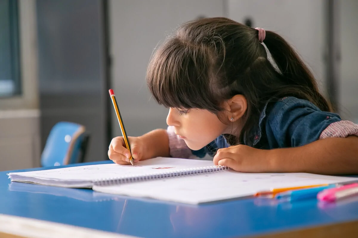 Menina concentrada escrevendo em um caderno com um lápis amarelo, rodeada de canetas coloridas em um ambiente escolar.