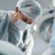 Profissional de saúde com jaleco, máscara e touca cirúrgica realiza mapeamento cerebral intraoperatório em quirófano, paciente acordado.