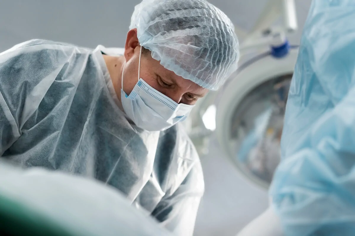 Profissional de saúde com jaleco, máscara e touca cirúrgica realiza mapeamento cerebral intraoperatório em sala de cirurgia, paciente acordado.
