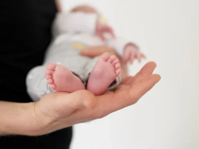 Mão de adulto segurando um bebê, foco em seus pés rosados e dedos definidos, iluminados com luz suave.