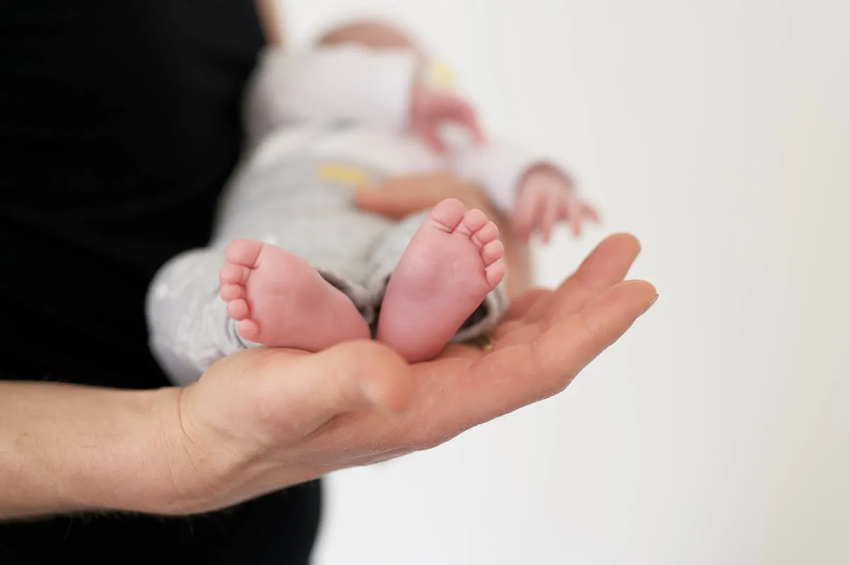 Mão de adulto segurando um bebê, foco nos pés rosados e dedos definidos, iluminados com luz suave.