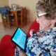 Pessoa idosa sentada em um sofá vermelho com tablet, blusa floral e óculos; logomarca da NeuronUP e gráficos de envelhecimento ativo.