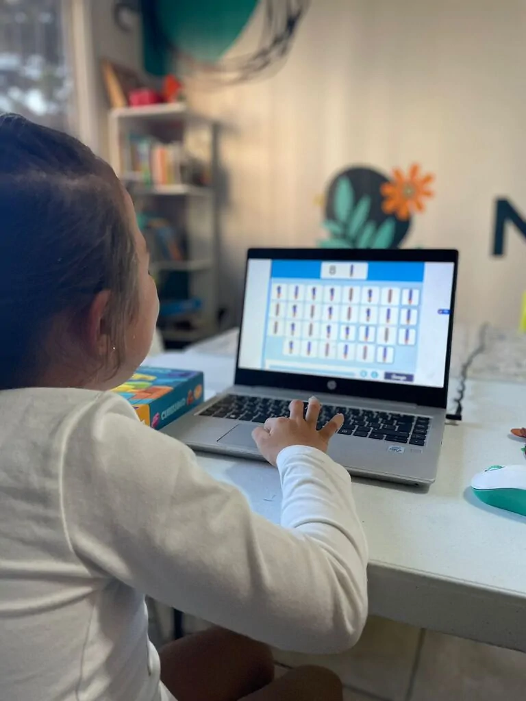 Menina interage com NeuronUP no Brainkare diante de um laptop; a tela mostra uma grade e ambiente com estantes.