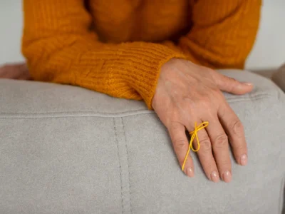 Mão repousando sobre um sofá claro, com suéter laranja e laço amarelo no pulso, cena calma para EM.