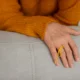 Mão repousando sobre um sofá claro, com suéter laranja e laço amarelo no pulso, cena calma para EM.