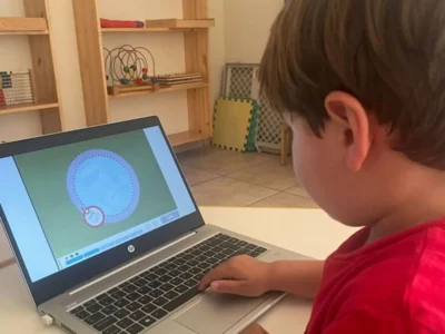 Criança sentada em frente a um laptop no Brainkare, usando NeuronUP; a tela mostra uma interface educativa e estantes com brinquedos ao fundo.