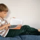 Menino lendo um livro em um sofá, camiseta listrada e ambiente minimalista; foco em consciência fonológica.