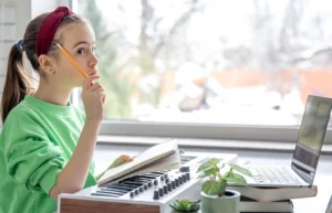 Menina sentada em frente a uma mesa com lápis, teclado musical e laptop; a janela mostra uma paisagem de inverno ao fundo, ambiente de neurodidática musical.