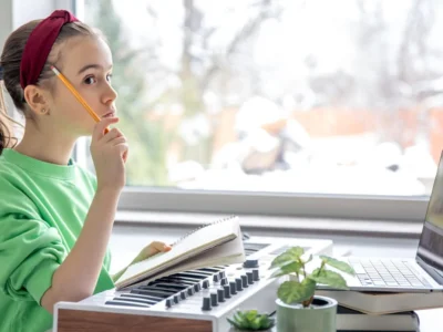 Menina sentada em frente a uma mesa com lápis, teclado musical e laptop; a janela mostra uma paisagem de inverno ao fundo, ambiente de neurodidática musical.