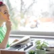 Menina sentada em frente a uma mesa com lápis, teclado musical e laptop; a janela mostra uma paisagem de inverno ao fundo, ambiente de neurodidática musical.