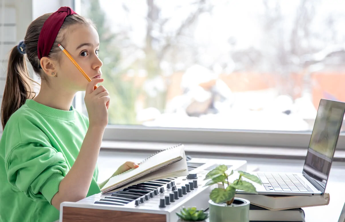 Menina sentada diante de uma escrivaninha com lápis, teclado musical e laptop; a janela mostra uma paisagem invernal ao fundo, ambiente de neurodidática musical.