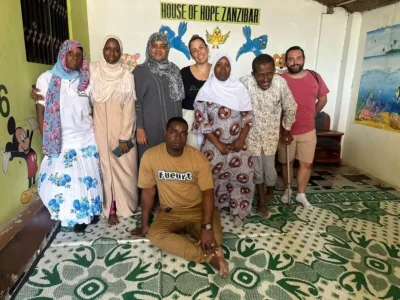 Grupo de voluntários da PsicoNED e da Fundação NED em Zanzibar, em pé e sentados em uma sala com mural 'HOUSE OF HOPE ZANZIBAR' e desenhos de peixes, sorridentes.