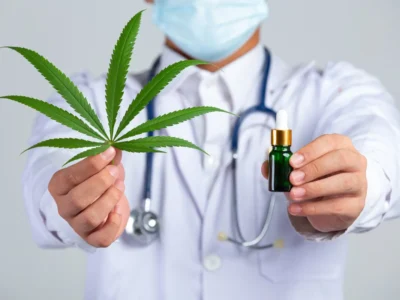 Profissional de saúde com jaleco branco e máscara segura uma folha de cannabis e uma garrafa de tintura; fundo neutro.