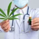 Profissional de saúde com jaleco branco e máscara segura uma folha de cannabis e uma garrafa de tintura; fundo neutro.