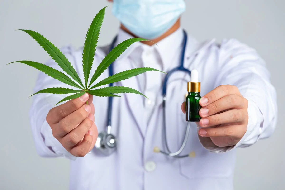 Profissional de saúde com jaleco branco e máscara segura uma folha de cannabis e uma garrafa de tintura; fundo neutro.