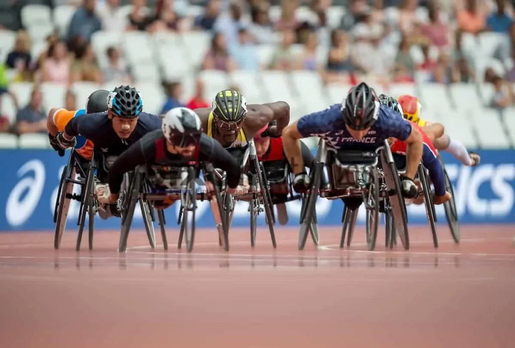 Grupo de atletas paralímpicos em uma corrida de cadeira de rodas, concentrados rumo à linha de chegada em um estádio.