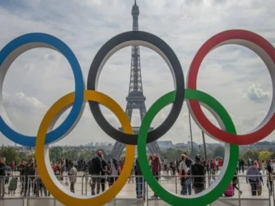Cinco anéis olímpicos em primeiro plano diante da Torre Eiffel, com pessoas ao redor em um ambiente esportivo festivo.
