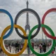 Cinco anéis olímpicos em primeiro plano diante da Torre Eiffel, com pessoas ao redor em um ambiente esportivo festivo.