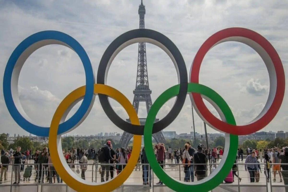 Cinco anéis olímpicos em primeiro plano diante da Torre Eiffel, com pessoas ao redor em um ambiente esportivo festivo.