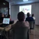 Grupo de participantes em uma sala de formação observando uma projeção com círculos coloridos e números, com um laptop sobre a mesa e luz natural.