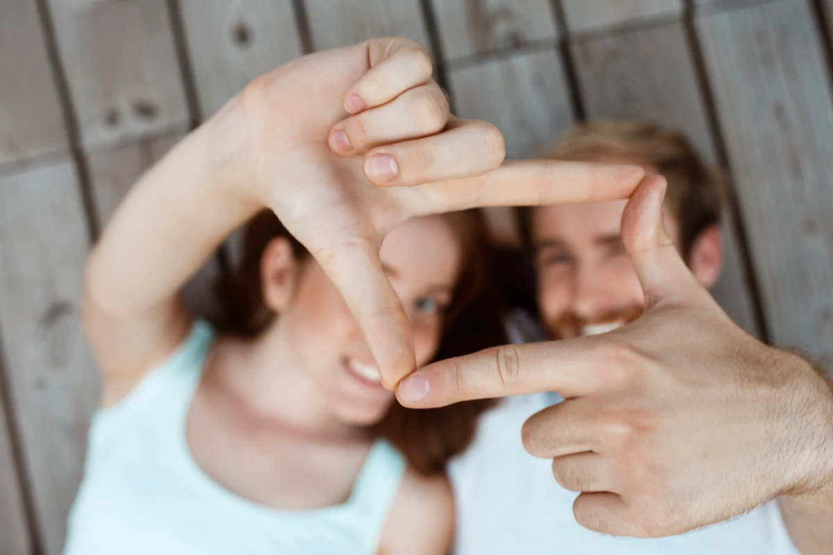 Duas pessoas deitadas sobre uma superfície de madeira, sorrindo; uma delas faz moldura com as mãos.