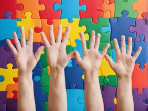 Várias mãos de diferentes idades erguidas diante de um fundo colorido com peças de quebra-cabeça, simbolizando diversidade e inclusão.