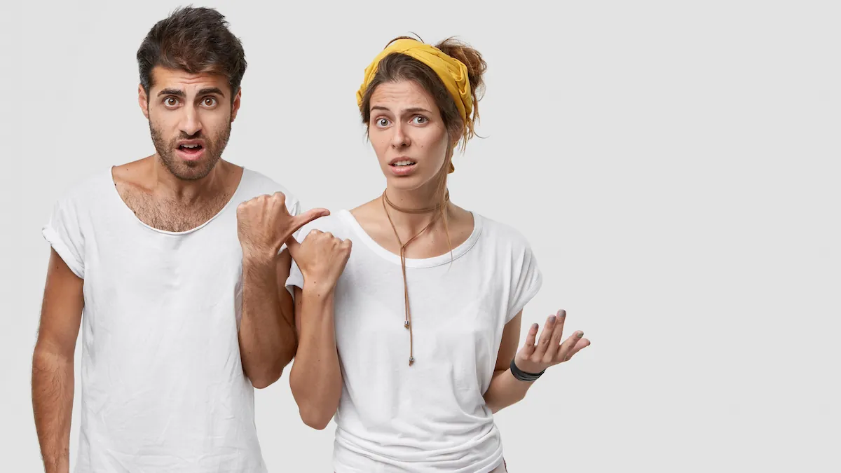 Homem de cabelo escuro e barba aponta para mulher de coque castanho, faixa amarela, em camiseta branca; fundo cinza.
