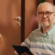 Homem idoso sentado com uma camisa listrada segura um tablet; fundo terracota, lâmpada exposta e plantas, iluminação quente.