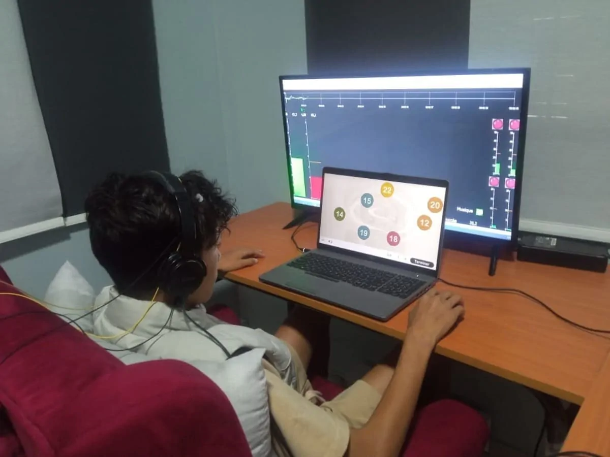 Pessoa sentada em cadeira, fones de ouvido, diante de laptop e tela com gráficos; ambiente claro e tranquilo.