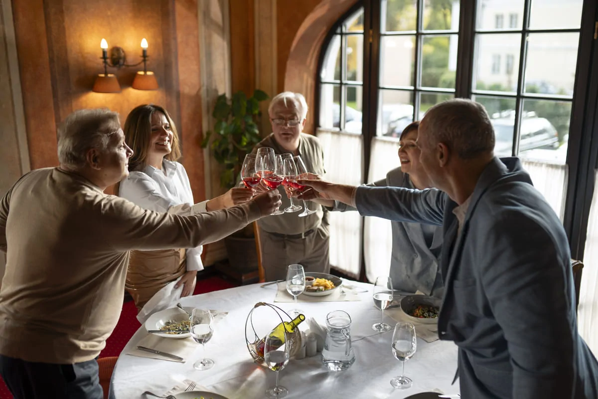 Grupo de cinco pessoas brindando em restaurante elegante, mesa posta com pratos e copos cheios, iluminação natural suave.