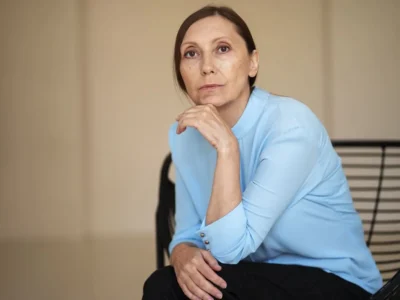 Mulher sentada em uma cadeira moderna com blusa azul-claro, expressão séria; fundo neutro e iluminação suave.