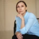 Mulher sentada em uma cadeira moderna com blusa azul-claro, expressão séria; fundo neutro e iluminação suave.