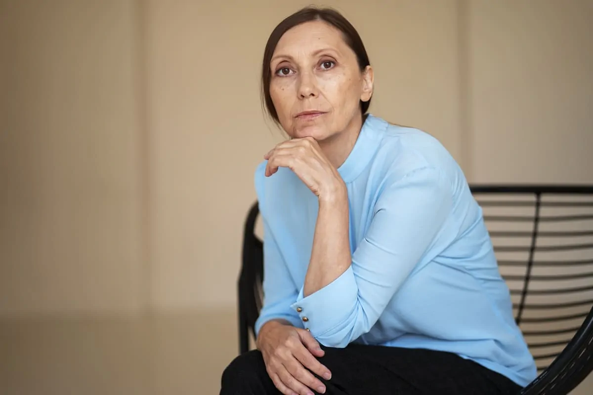 Mulher sentada em cadeira, cabelos longos escuros, camisa azul; expressão séria e pensativa com a mão no queixo, fundo neutro.