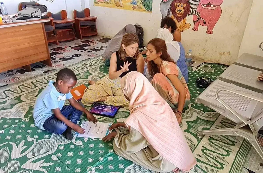 Criança negra sentada no chão de sala educativa, concentrada em papéis; duas mulheres orientam, uma observa, ao fundo mesas.