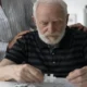 Homem idoso concentrado resolvendo um quebra-cabeça em um espaço interno, com apoio próximo de outra pessoa.