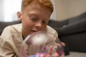 Menino ruivo deitado no chão observa uma esfera luminosa com luzes coloridas, em ambiente neutro.