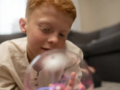 Menino com cabelo ruivo deitado no chão olhando para uma esfera luminosa; sala de estar ao fundo com sofá e brinquedos.