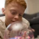 Menino com cabelo ruivo deitado no chão olhando para uma esfera luminosa; sala de estar ao fundo com sofá e brinquedos.