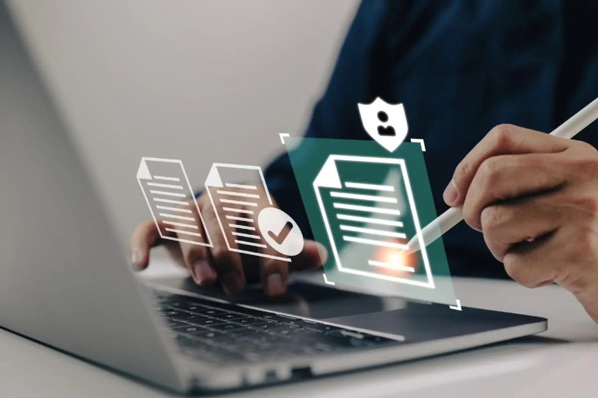 Pessoa digitando em um laptop, com ícones de documentos e verificação ao lado; caneta na tela em ambiente moderno.