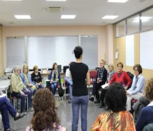Mulher em pé, de frente para grupo sentado em círculo numa sala iluminada, com mesas ao fundo e materiais visíveis.