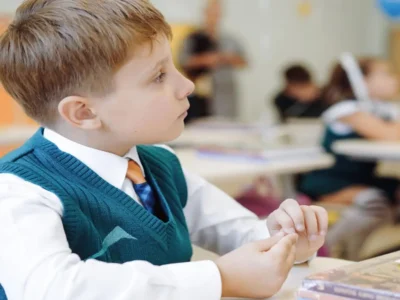 Criança em sala de aula atenta veste uma colete verde, camisa branca e gravata; fundo com outros estudantes.