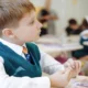 Criança em sala de aula atenta veste uma colete verde, camisa branca e gravata; fundo com outros estudantes.