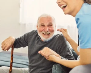 Homem idoso sorrindo em um sofá, acompanhado por uma pessoa mais jovem que segura a mão dele, em um ambiente acolhedor.