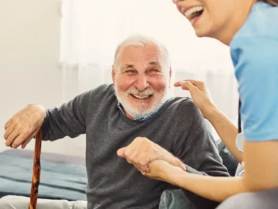 Homem idoso sorrindo em um sofá, acompanhado por uma pessoa mais jovem que segura a mão dele, em um ambiente acolhedor.