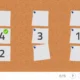 Interface tipo painel de cortiça com três folhas bege, números 1 e 2, botão Pronto, contador 0/5 e ícones de coração e reprodução.