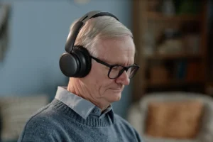 Homem idoso com fones de ouvido, sentado e concentrado, suéter cinza e camisa azul, fundo com estantes e sofá.