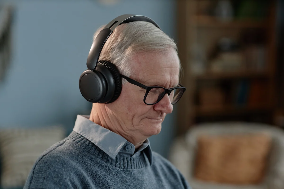 Homem idoso com fones de ouvido, sentado e concentrado, suéter cinza e camisa azul, fundo com estantes e sofá.