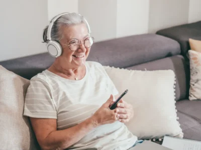 Mulher idosa sentada em um sofá com fones de ouvido e celular, sorrindo em um ambiente confortável e acolhedor.