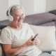 Mulher idosa sentada em um sofá com fones de ouvido e celular, sorrindo em um ambiente confortável e acolhedor.